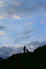 Abendspaziergang mit Hund. Silhouette einer Frau mit Hund auf einem Berg vor Wolkenhimmel