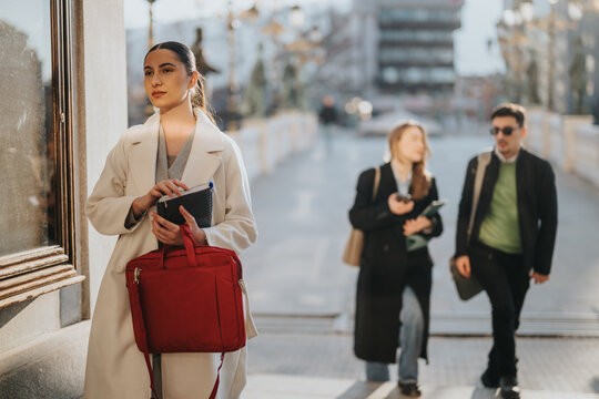 A stylish woman stands confidently on a city street carrying a red bag, with blurred figures of colleagues in motion around her, depicting modern entrepreneurship, teamwork, and aspirational dynamics.