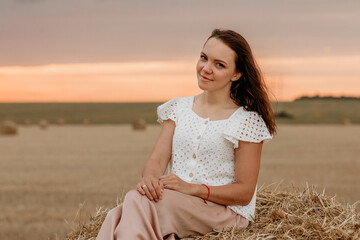Beautiful young brunette girl in a field of haystacks at sunset in summer