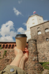Brown paper cup of natural coffee in women's hand on against backdrop of Olaf's Tower in city Vyborg in blur, Russia on sunny day