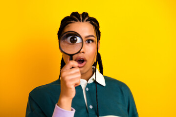 Young woman holding magnifying glass with playful expression in front of vibrant yellow backdrop...
