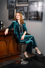 Elegant mature woman in teal velvet suit sitting at bar counter with cup of coffee in hand...