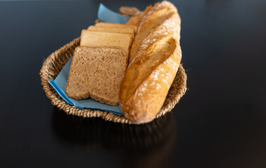 Whole baguette and sliced toast bread—both whole grain and white—arranged on blue napkins in a wicker basket. Placed on a black table for a cozy, rustic bakery vibe