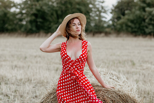 Beautiful young girl in a red polka dot dress and hat in a field of haystacks