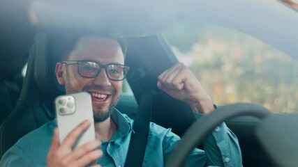 Excited man in glasses raising fist while smiling inside parked vehicle. Celebrating good news after reading message on smartphone. Expressing joy and satisfaction during cheerful moment. - Powered by Adobe