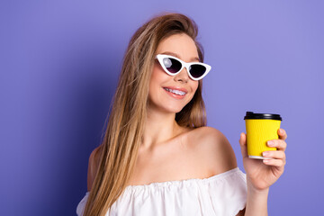 Obraz premium Chic young woman wearing stylish sunglass holding a yellow coffee cup against purple background, expressing joy and charm