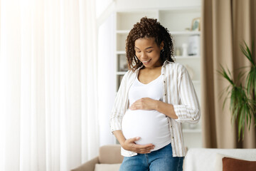 Black pregnant woman looking down at belly and smiling while standing near window. Intimate and...