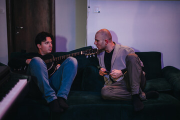 Two men are discussing music ideas on a sofa, with one holding an acoustic guitar. The atmosphere...