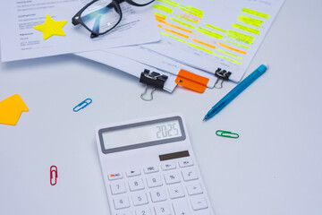 Finance and budget planning concept with eyeglasses, a calculator displaying 2025. Documents with highlighted text, and colorful office supplies on a white desk.