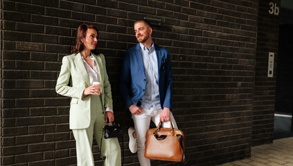 Two young business professionals, a man and a woman, are standing by a brick wall, enjoying a coffee break after work, holding their work bags and engaging in conversation