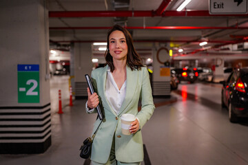 Businesswoman walking through a parking garage after work, carrying a laptop and takeaway coffee, heading home or to another meeting, exemplifying a modern, ambitious lifestyle