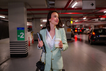Young businesswoman walking through a parking garage after work, holding a laptop under one arm and a takeaway coffee, confidently heading towards her car in the evening