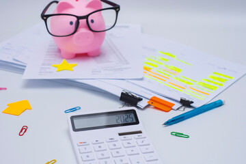 Finance and budget planning concept with a pink piggy bank wearing eyeglasses, a calculator displaying 2025. Documents with highlighted text, and colorful office supplies on a white desk.