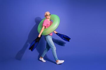Cheerful senior woman wearing a striped t-shirt and blue jeans, holding swimming flippers and a...