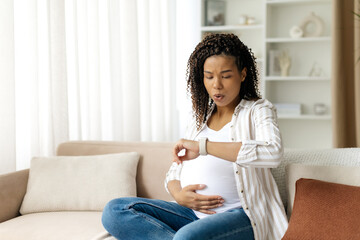Black pregnant woman looking at smartwatch and breathing calmly, counting contractions. Represents...