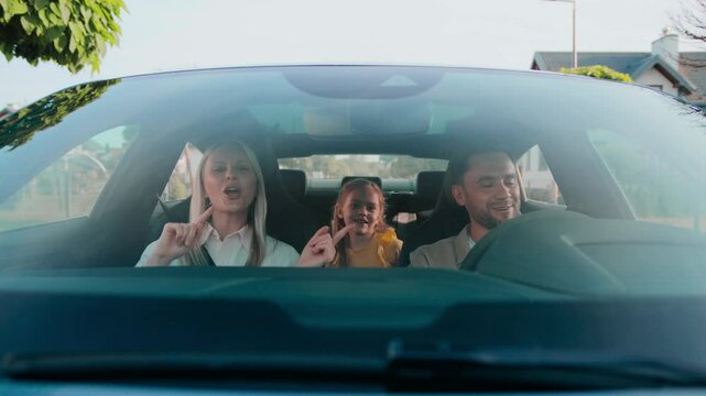 Excited woman singing song while sitting in passenger seat. Bearded man smiling and glancing toward her. Little girl in yellow holding teddy bear dancing in backseat. Enjoying upbeat music together.