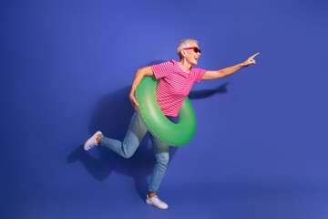 Joyful senior woman posing in stylish outfit with a green inflatable ring against a vibrant purple...