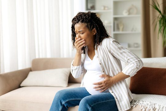 Black pregnant woman covering mouth in discomfort, experiencing nausea. Realistic moment of morning sickness or digestive issue in cozy home environment, copy space