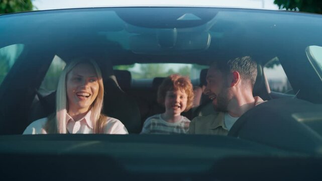 Laughing woman in white shirt enjoying funny moment during car ride. Curly-haired child holding teddy bear with mouth open in excitement. Smiling man turning head toward backseat sharing joy.