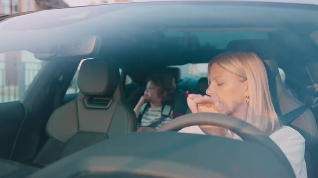 Serious blonde woman holding phone and reading message while sitting behind steering wheel. Concentrating on screen. Thoughtful boy in car seat resting head on hand and gazing through window silently.