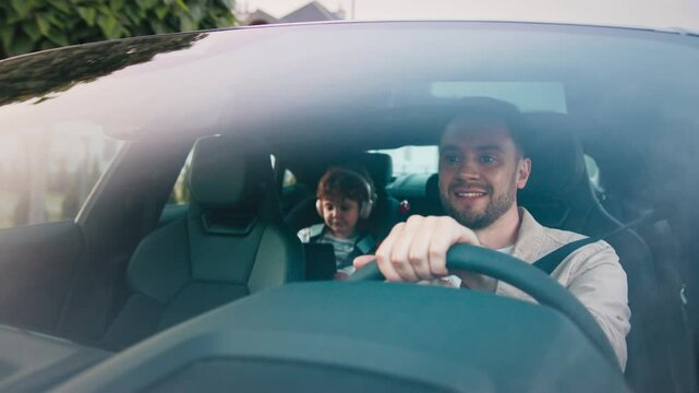 Focused man turning head slightly while steering wheel and cruising through neighborhood. Curious curly-haired boy in car seat wearing headphones and watching video on tablet during relaxed drive. - Powered by Adobe
