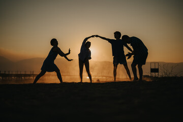 A group of four silhouetted friends creates artistic poses at sunset on a beach.
