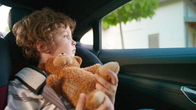 Curly-haired boy resting quietly in car seat holding brown teddy bear against chest. Gazing through window with calm. Enjoying peaceful drive past suburban homes in afternoon light.