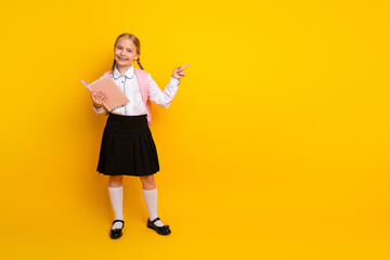 Cheerful schoolgirl with book and braids posing on a yellow background ready for school and new...
