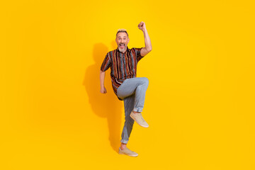 Joyful mature man with blond hair in stylish attire leaping against a bright yellow background...