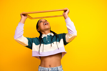 Playful modern young woman holding a wooden frame smiling against bright yellow background...