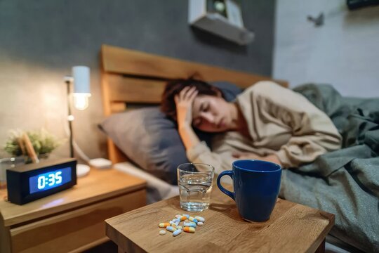 Person lying in bed, holding their head, looking distressed. glass of water and cup are on bedside table with pills. clock shows 035, creating sense of urgency and concern