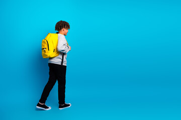 Young student wearing a yellow backpack walking against a blue background in casual grey outfit...