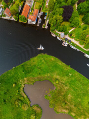 A serene riverside scene with houses, boats, and a single sailboat gliding through the water. A...