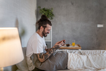 Happy tattooed hipster man using smartphone in bed after waking up