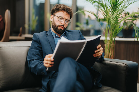 A businessman attentively reading through detailed documents while seated on a comfortable couch in a contemporary office. This image captures professionalism, focus, and the corporate environment.