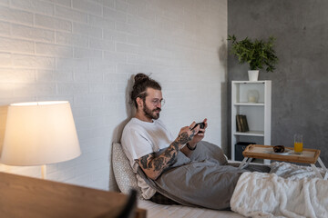 Young man relaxing in bed using smartphone in modern apartment