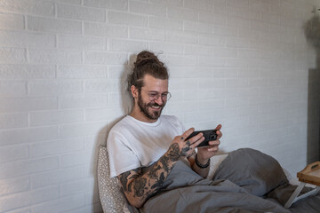 Happy hipster man playing mobile video game in bed