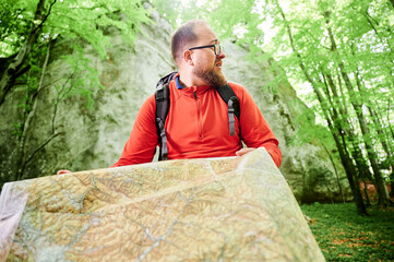 Close up of bearded tourist man studies map while stands in front of large rock in dense forest....