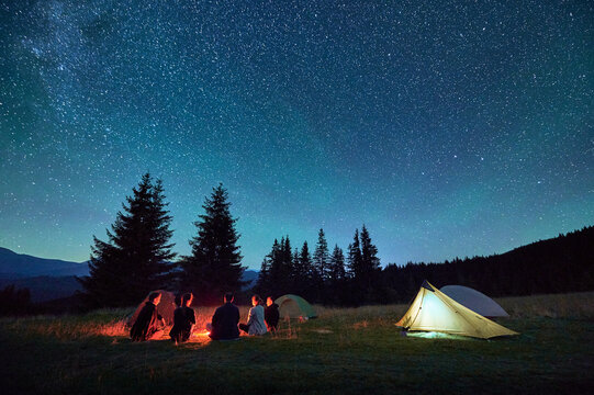 Group of friends hikers resting around campfire, surrounded by illuminated tents under stunning starry sky. Milky Way stretches above, creating magical atmosphere in peaceful mountain wilderness.