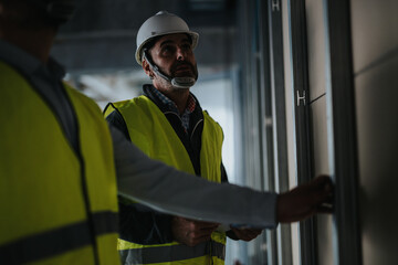 Two construction professionals wearing protective equipment and high-visibility vests evaluate...