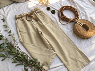 Flat lay of beige linen wide-leg pants on a white cotton sheet with eucalyptus branches, a woven belt, and soft sunlight, creating a boho minimalist fashion scene.
