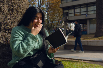 young woman reading a book smiling happily with one hand on her mouth