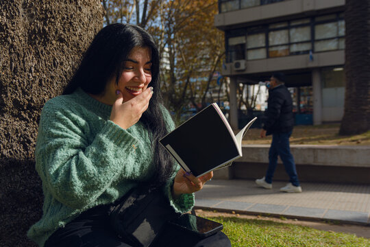 young woman reading a book smiling happily with one hand on her mouth