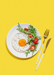 Fried eggs with arugula and cherry tomatoes on a white plate on a bright yellow background