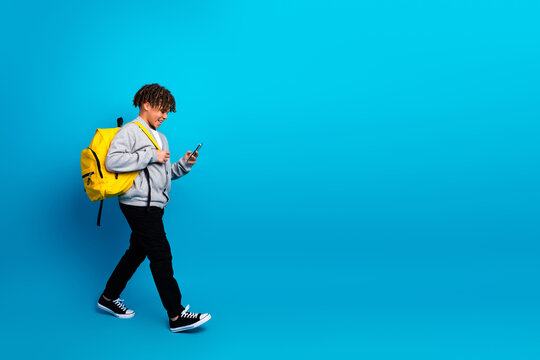 Smiling schoolboy with yellow backpack walking while looking at smartphone against a vibrant blue background - Powered by Adobe