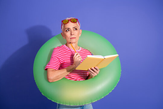 A stylish senior woman with short gray hair reading a book while wearing a green inflatable ring against a purple background