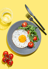 Fried eggs with arugula and cherry tomatoes on a dark plate on a bright yellow background