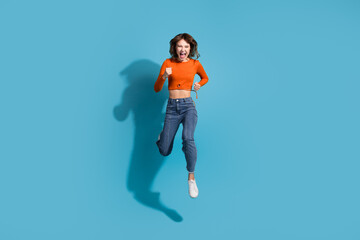 Joyful young woman in stylish orange top and jeans leaping passionately in front of a vibrant blue...