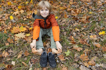 cheerful child takes off his shoes and sits barefoot among autumn leaves on a warm autumn day. Joy,...