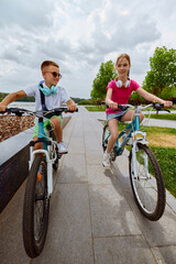 Wheels and words. Kids cycle along a picturesque park trail beneath a cloudy sky, embodying the...